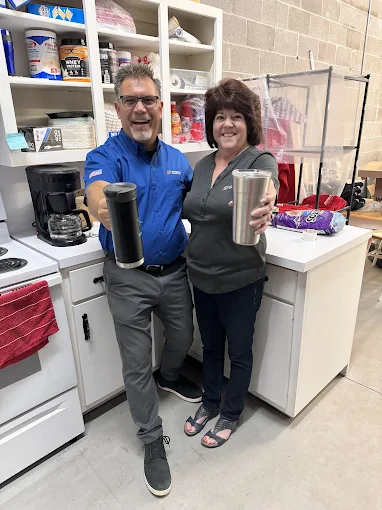 Two people standing in a break room, smiling and holding up travel mugs. Shelves with snacks and a coffee maker are visible in the background.