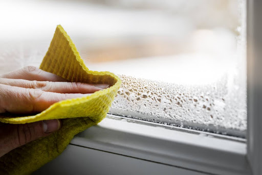 A close-up of a hand using a cloth to wipe condensation from a window due to high humidity in a home.