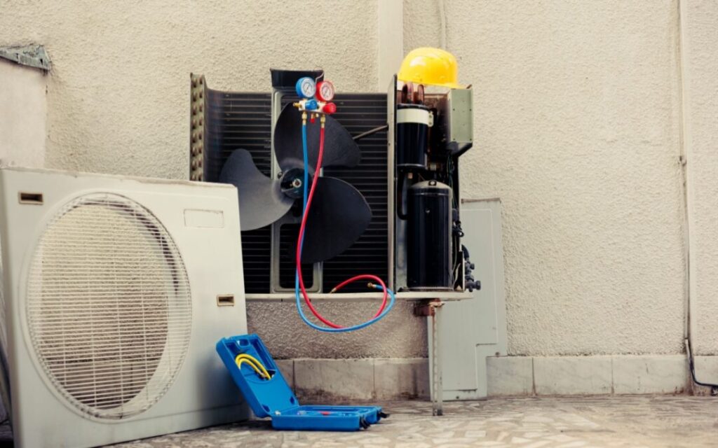 An open outdoor air conditioning unit with attached gauges, a yellow hard hat, and a blue tool case on the ground next to a tiled surface.
