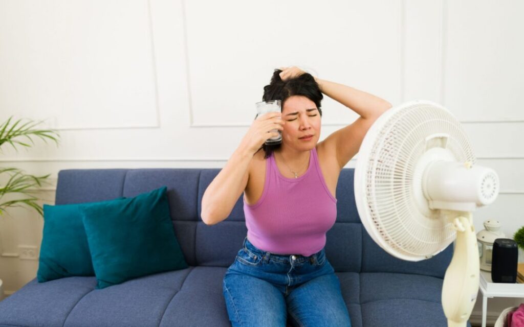A woman sitting on a couch holds an ice pack to her head and sits in front of a fan, appearing overheated.