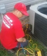 A technician in a red shirt and cap is servicing an outdoor air conditioning unit using various diagnostic tools and cables.