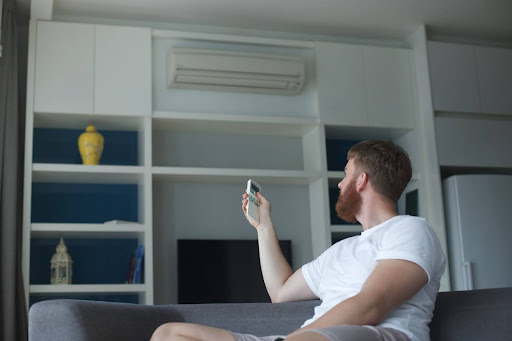 A man sitting on a couch points a remote control at an air conditioner mounted on the wall in a modern living room.